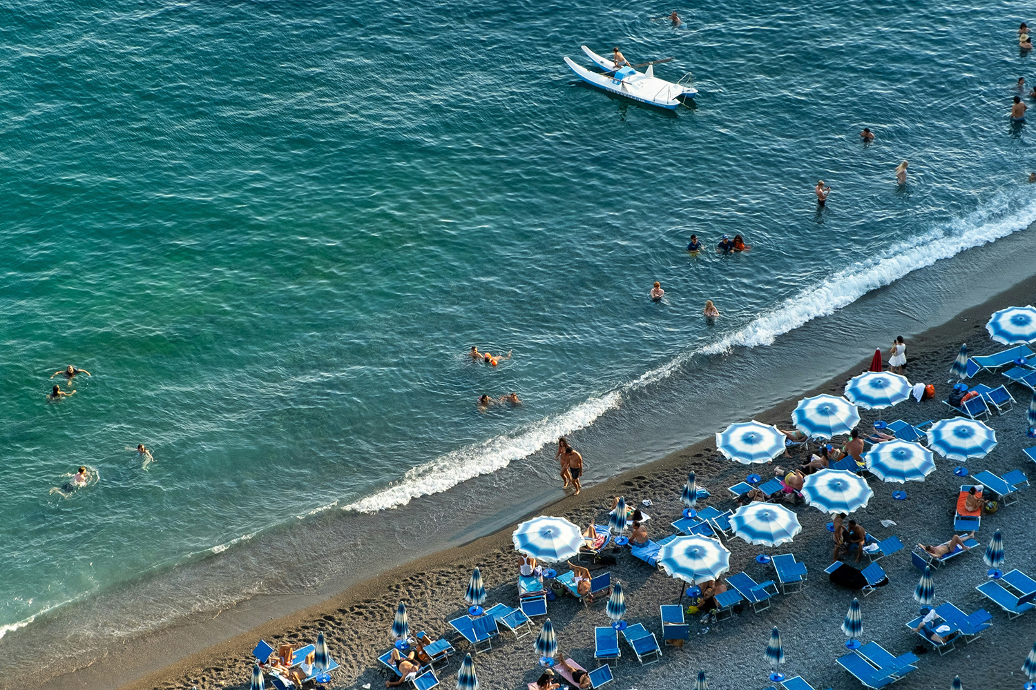 Swimmers in clear turquoise water beside a pebble beach with blue umbrellas on the Amalfi Coast.