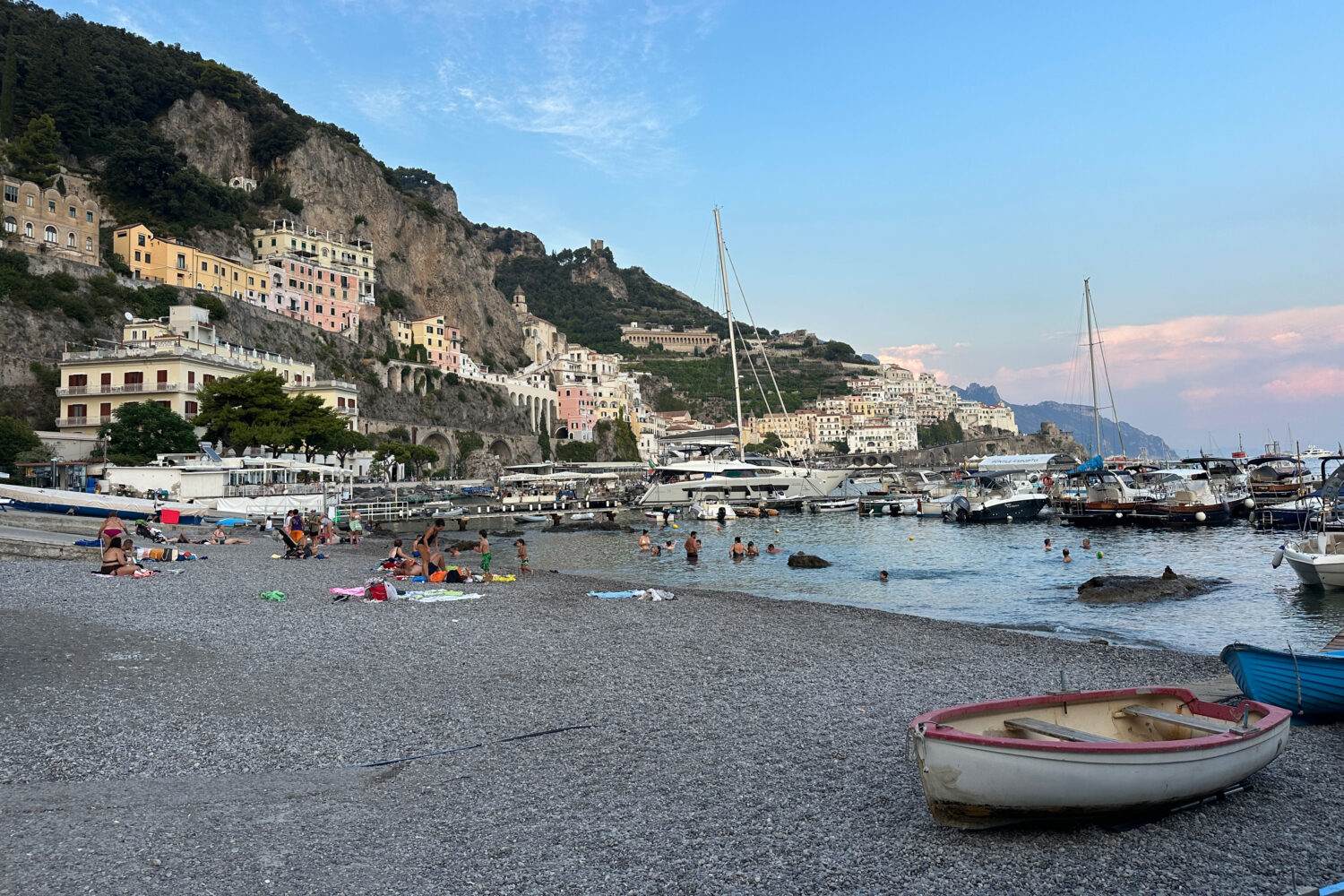Families and locals swimming at a small public beach beside Amalfi's marina, with boats, colourful buildings and coastal mountains in the background.