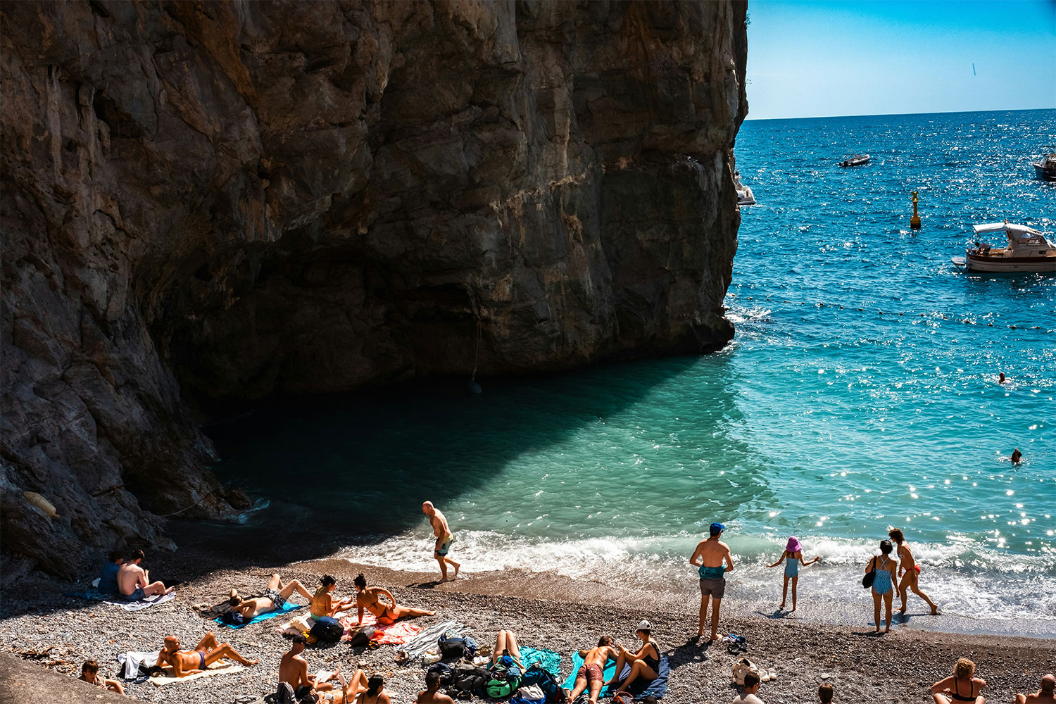 Beachgoers on a pebble shore beneath a tall cliff, with swimmers in turquoise water and boats offshore.