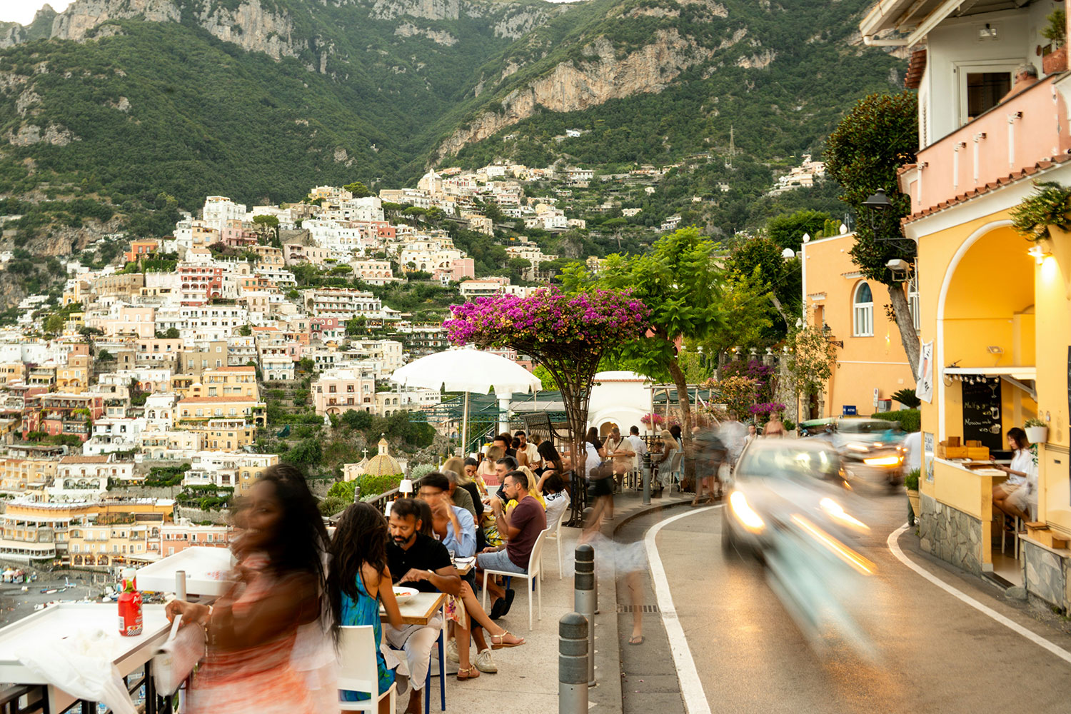Cars passing beside outdoor restaurant tables on a narrow road in Positano