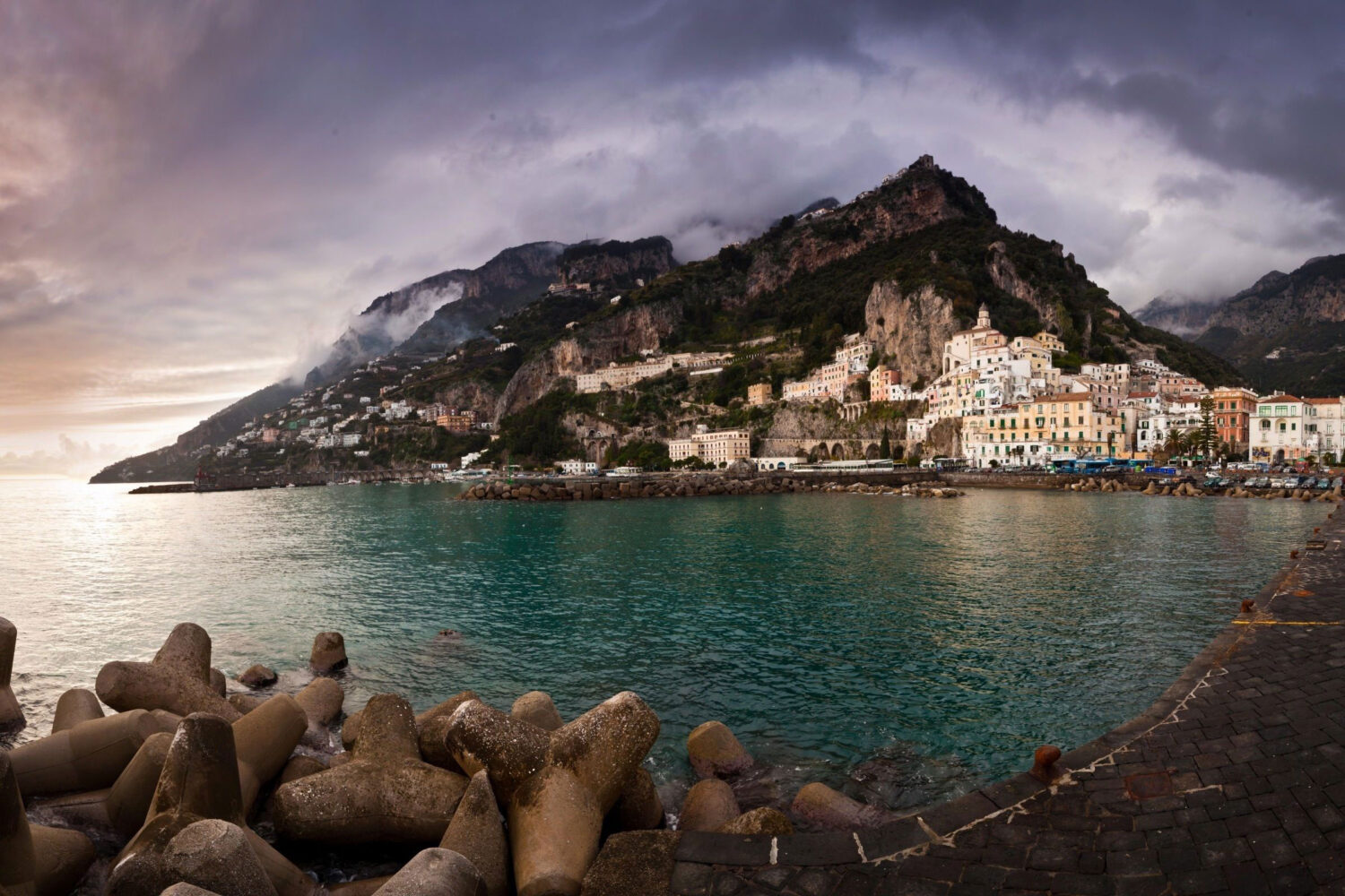 Amalfi waterfront beneath low clouds over the mountains, with turquoise water and harbour breakwaters.