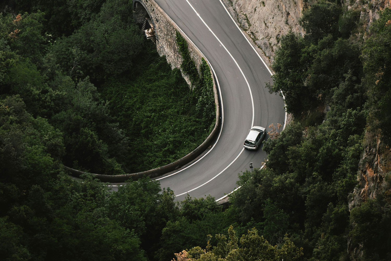 Two cars driving along a winding mountain road surrounded by dense green trees on the Amalfi Coast.